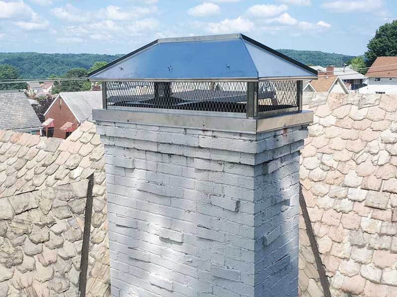 Close up of a stainless steel chimney cap on a white chimney sitting on wood shingle roofing and buildings in the background - trees and blue sky with clouds also in the background