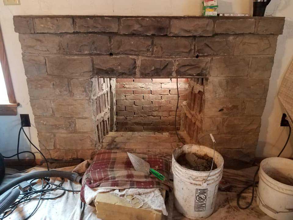  before restoration of PriorFire firebox custom size built out of fire brick. with chiseled rock surround and mantle - light in firebox - mortar and cleaning supplies in the foreground.