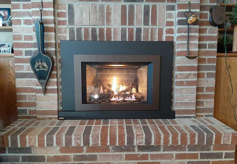 Wood fireplace insert with black and brown trim and fire burning - tools on the left and right with a brick hearth and surround.