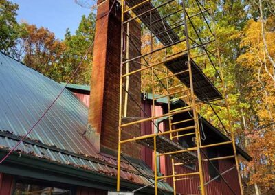 Masonry chimney before repair scaffolding up the side of a red wooden home with green metal roof and fall trees in the background.