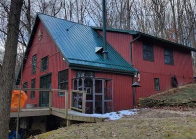Chimney after repair replaced with a black pipe on red wooden home with green metal roof with winter trees in the background.