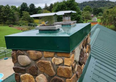 Beautiful new stainless steel chase cover green with a green metal roof. Chimney chimney is made of large river rock and there are trees and a cloudy sky in the background.