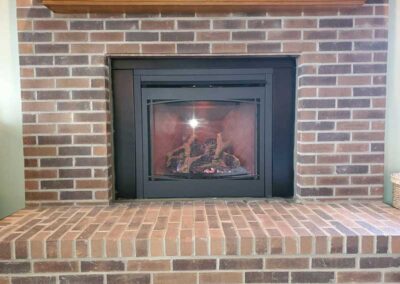 New gas fireplace insert with red brick surround and hearth with wooden mantel.