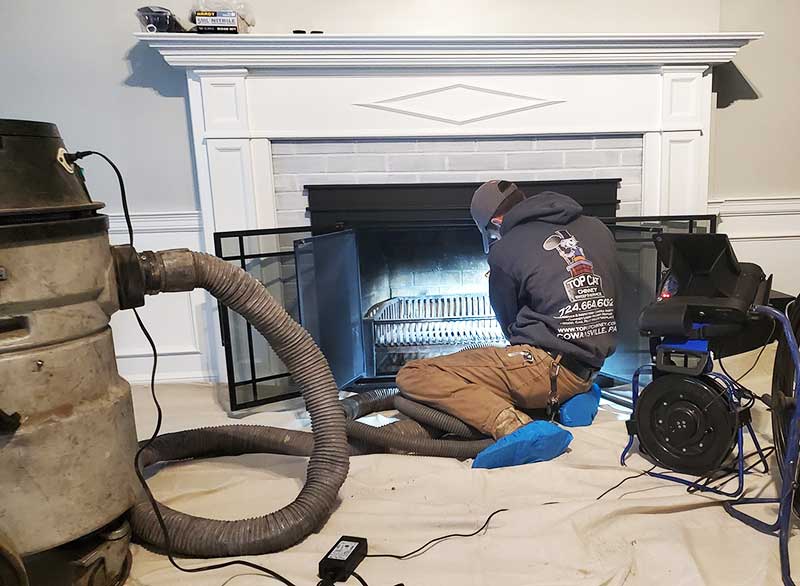 Technician cleaning a fireplace wearing Top Cat Logo sweatshirt and hat.  He is wearing bootie covering on his shoes and has a tarp on the floor.  To the left is a vacuum.