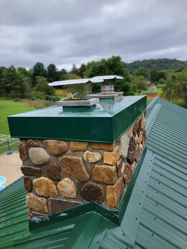 New stainless steel chimney cap painted green to match the green metal roofing - cloudy skies and trees in the background - the chimney is made of irregular river rock