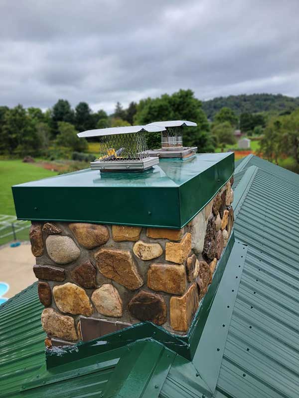 Stock image of chase cover after replacement - Top Cat Chimney - Cowansville, PA Beautiful new stainless steel chase cover green with a green metal roof. Chimney chimney is made of large river rock and there are trees and a cloudy sky in the background.