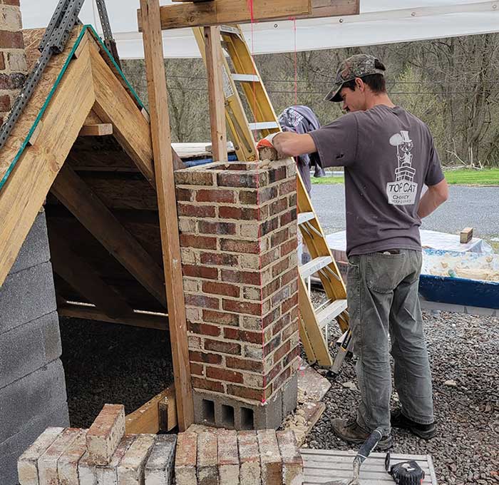 Tech on the ground starting new construction of chimney with a frame to the left and framing around it - He is wearing a gray Top Cat logo t-shirt.