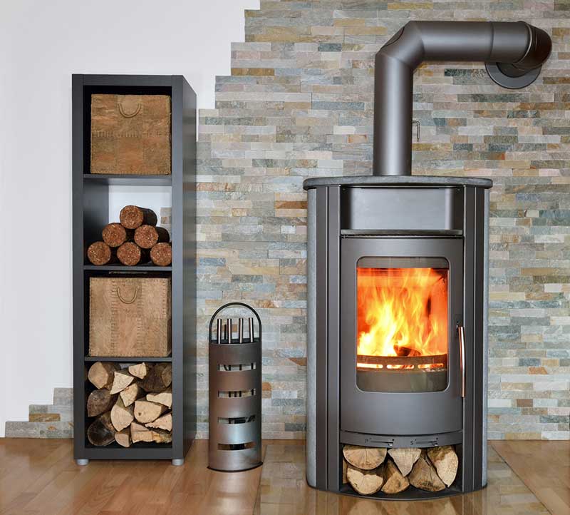 Modern tall wood stove with pipe going out the back wall and wood storage underneath.  Shelving to the left with wood storage and tools in the middle.