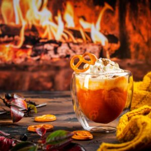 an orange beverage in a clear mug with whipped topping settled in front of a blazing fireplace